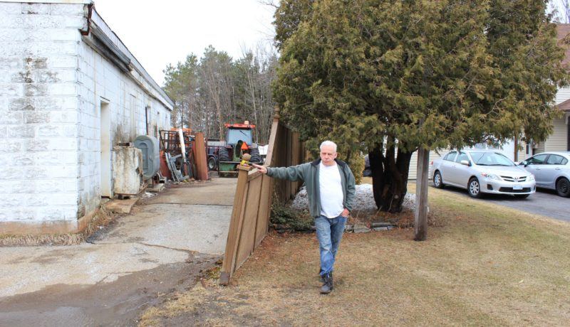 Yves Frappier standing between the properties. We can clearly see how close the two properties are to each other. On his side is zoned residential and on the other side of the fence is zoned industrial.